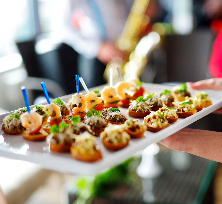 Un vassoio bianco viene servito, con tartine colorate e spiedini di gamberi in primo piano.